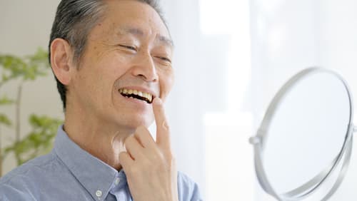 Elderly man examining his teeth in the mirror with a smile, showcasing the confidence and function of wearing dentures.